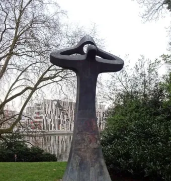 Der Mahner, a tall abstract sculpture with raised arms, stands on a lawn beside water and modern buildings.