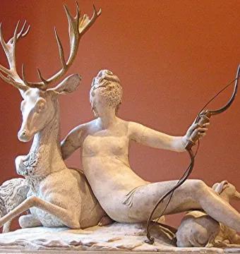 Marble sculpture of Diana seated with a bow, a large deer, and two hounds on a stone base.