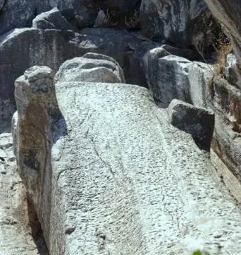 Unfinished Kouros of Apollonas carved from a rock face in an ancient quarry, with a rough, pockmarked surface and surrounding dark stone.