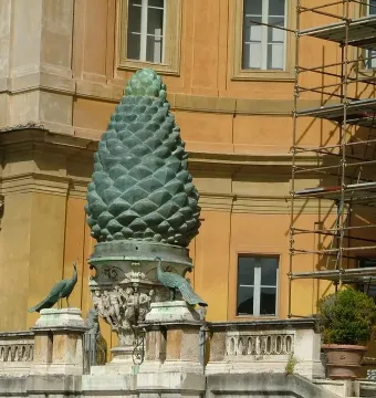La Pigna bronze pine cone sculpture with peacocks on an ornate stone balustrade, in front of a building partly covered by scaffolding.