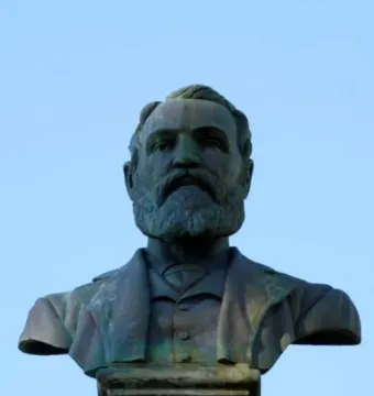 Weathered bronze bust of Maitland Brown in a suit and tie on a stone pedestal against a blue sky.