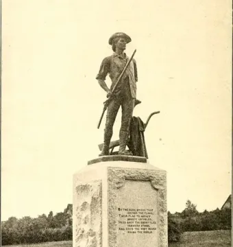 The Minute Man statue stands on a stone pedestal in a grassy area, holding a long gun with carved reliefs and inscriptions on the base.