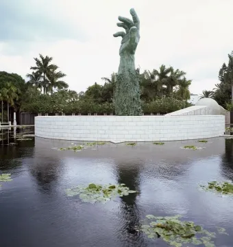 Holocaust Memorial sculpture of a towering bronze hand formed by human figures, rising beside a white curved wall and reflecting pool with palm trees nearby.