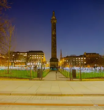 The Melville Monument rises above glowing lights in St Andrews Square at twilight, with illuminated buildings in the background.