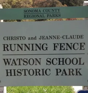 Light-green and white historical site sign for Running Fence on a grassy lawn with trees behind it.