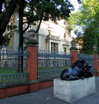 Dark metal guarding lion sculpture on a light stone pedestal outside a fenced building.