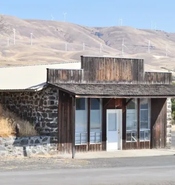 Rustic building at the entrance to Maryhill Stonehenge with wind turbines on the hills behind it.