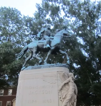 Stonewall Jackson equestrian statue on a stone pedestal with trees and a building behind it.