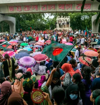 Anti Terrorism Raju Memorial Sculpture beside a large crowd with umbrellas, Bangladeshi flags, and a concrete archway in the background.