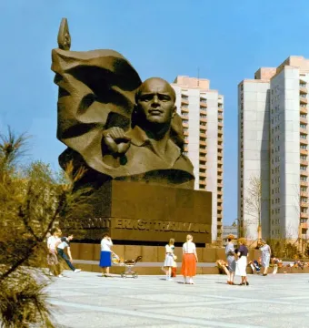 Ernst Thälmann Memorial in a wide public square with apartment buildings behind it and people walking nearby.