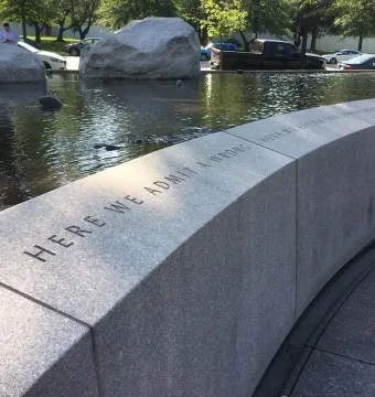 Curved stone wall of the Japanese American Memorial inscribed with “HERE WE ADMIT A WRONG,” beside a reflecting pool and boulders.