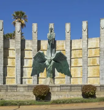 Monumento a Franco with a bronze robed figure holding a sword atop a winged figure in front of a curved stone wall and pillars.