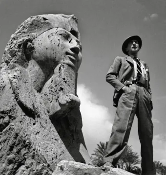 The Sphinx of Memphis with a man in a suit and fedora standing beside it under a cloudy sky.
