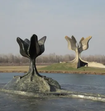 A dark Stone Flower sculpture in the foreground with the larger Kameni cvijet monument across the water on a grassy mound.