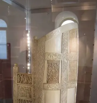 Throne of Maximian with a high arched back and ornate relief carvings inside a glass case.