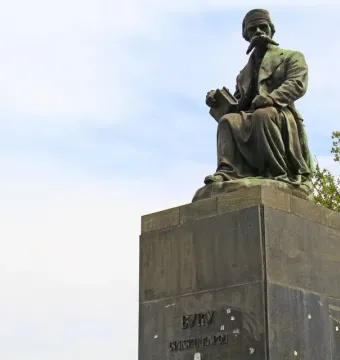 Bronze monument of Vuk Karadžić seated on a tall stone pedestal, holding a book, with trees and a cloudy sky behind it.