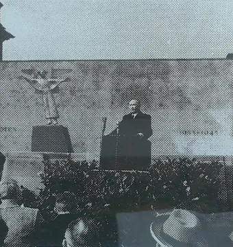 The Angel of Peace monument stands on a pedestal behind a man speaking at a podium to a crowd.