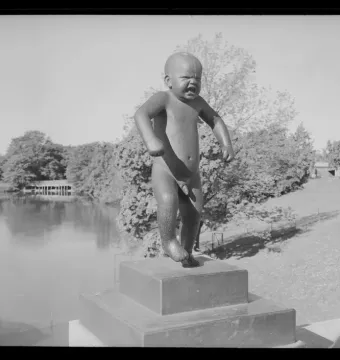 The Angry Boy sculpture stands on a pedestal outdoors beside water and trees.