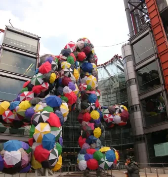 The Big 4 sculpture covered in colorful open umbrellas stands outside a modern glass building.
