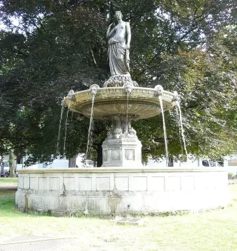 A classical draped female statue stands atop the Diana Fountain as water flows into a stone basin, with trees in the background.