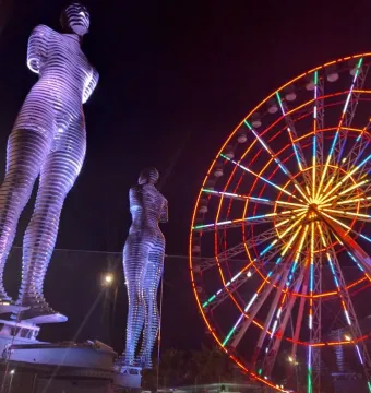 Ali and Nino statues lit in purple beside a brightly lit Ferris wheel at night.