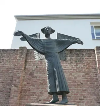 Angel of Peace statue with outstretched arms on a reddish stone pedestal, set against a brick wall and modern building.
