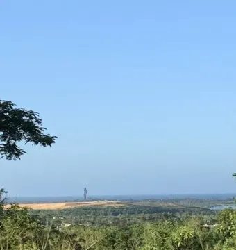 The Birth of the New World sculpture silhouetted against a clear blue sky, with trees in the foreground and water below.