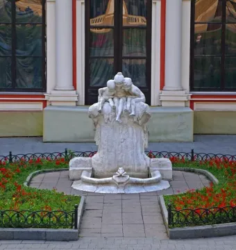 The Children and Frog sculpture on a weathered pillar, surrounded by a flowerbed with red blossoms and a low metal fence.