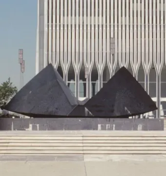 Cloud Fortress sculpture stands in a plaza before a tall ridged tower, with a spherical sculpture partly visible at right.