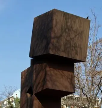 El Llarg Viatge, a tall rust-colored metal sculpture with stacked rectangular forms and tree shadows cast across it.