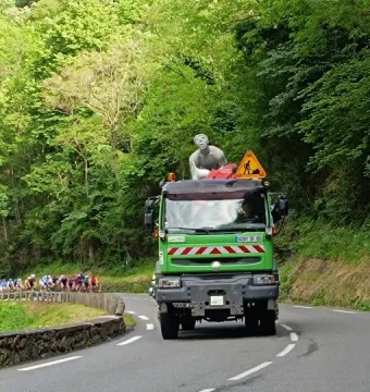 The Géant du Tourmalet statue mounted on a green utility truck on a winding mountain road, with cyclists in the distance.
