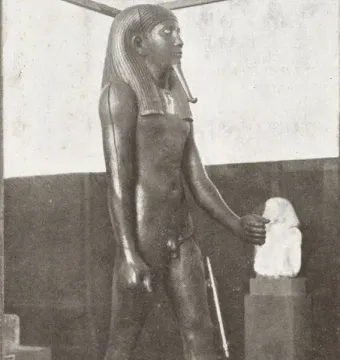 Statue of King Hor standing in a museum display, wearing a striped headdress and false beard and holding a staff.
