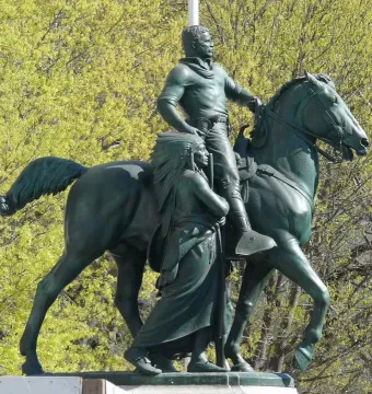Theodore Roosevelt Equestrian Statue with a horse, a walking figure in a feathered headdress, and leafy green trees behind it.