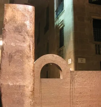 Topos V sculpture by Chillida with a tall pillar and an arched block against historic stone buildings.