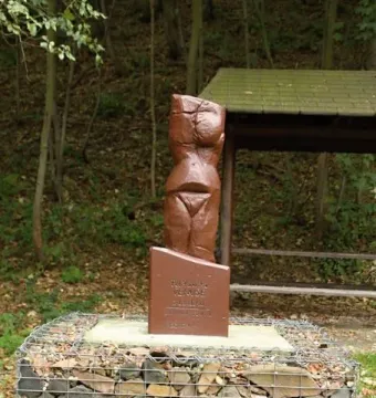 Venus of Petrkovice sculpture on an inscribed plinth and stone-filled gabion base in a wooded setting.