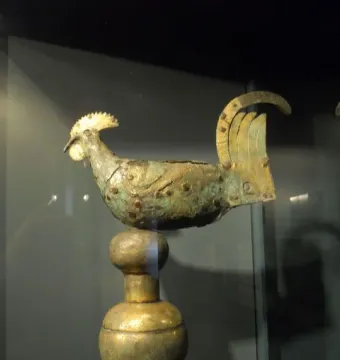 Gallo di Ramperto rooster sculpture with a weathered greenish-brown patina on a tall stand.