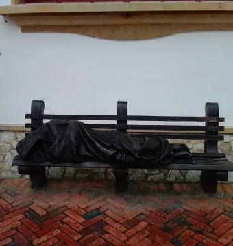 Jesus the Homeless sculpture of a draped figure lying on a park bench against a white wall.