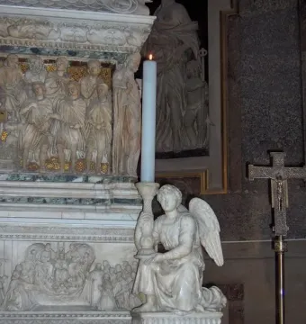 Kneeling white marble angel holding a tall candlestick with a lit candle, set before an ornate relief.