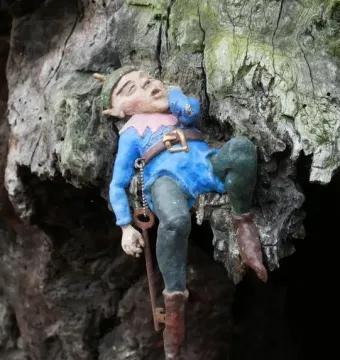 Sleeping gnome sculpture on the Elfin Oak, dressed in blue and green and holding a key against a weathered tree trunk.