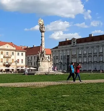 The Holy Trinity Monument stands in a sunny public square beside historic buildings and a grassy area with people walking nearby.