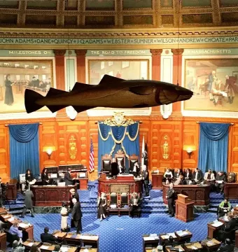 The Sacred Cod hangs above seated lawmakers in a wood-paneled legislative chamber.