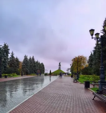 A paved park path leads toward the Salavat Yulaev Monument on a grassy mound, lined with trees and street lamps under an overcast sky.