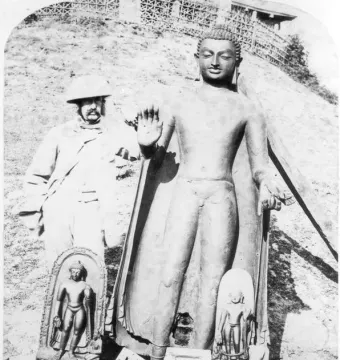 A man in colonial-era clothing and a pith helmet stands beside the Sultanganj Buddha, with two smaller carved stelae at the base.