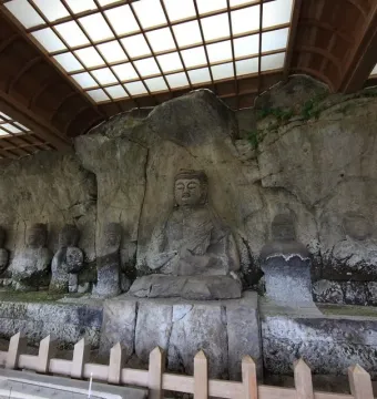 Usuki Stone Buddhas carved into a rock cliff beneath a protective wooden shelter with a fenced viewing area.