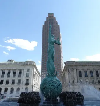 Fountain of Eternal Life sculpture rising from a plaza fountain, with water spraying around its base and city buildings behind it.