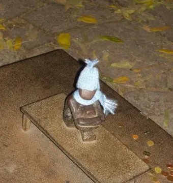 Järnpojke, a small dark sculpture of a boy wearing a light blue knitted hat and scarf on a tiny bench, with coins nearby.