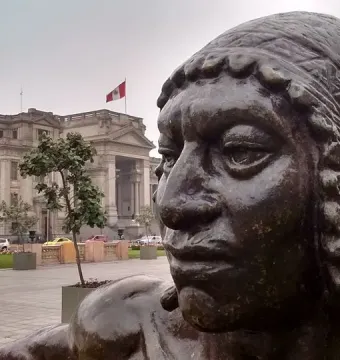Close-up of La Yunta bronze sculpture with a classical building and Peruvian flag in the background.
