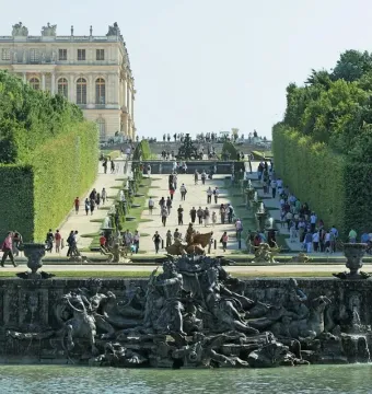 Neptune Fountain in Versailles with mythological sculptures and cascading water, set before a formal garden path leading toward the palace.
