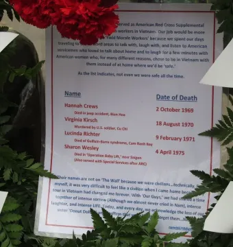 A laminated sign with names and text, framed by green leaves, white ribbons, and red flowers at the Vietnam Women\'s Memorial.