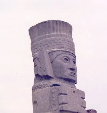 Tall stone Atlantean figure at Tula with an elaborate headdress and stylized face, seen against a bright sky.